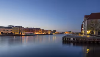 En lugn kvällsscen av en stadsbild vid vattnet. Upplysta byggnader kantar det lugna vattnet under en klarblå himmel som reflekterar levande ljus. En träbrygga sträcker sig in i floden, vilket ger djup till den fridfulla stadsvyn.