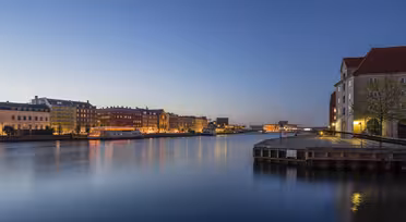 En lugn kvällsscen av en stadsbild vid vattnet. Upplysta byggnader kantar det lugna vattnet under en klarblå himmel som reflekterar levande ljus. En träbrygga sträcker sig in i floden, vilket ger djup till den fridfulla stadsvyn.
