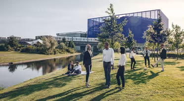 En grupp människor står och pratar på en gräsmatta nära en damm. I bakgrunden reflekterar en modern byggnad med glas- och metallstrukturer solljuset. Vissa människor sitter på gräsmattan, andra går i närheten.