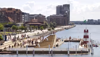 Människor njuter av en solig dag på en utomhusbadplats i en kuststad. Området har träplattformar, höga dykkonstruktioner och kantas av tegel och moderna byggnader. Vattnet är lugnt och en grön flagga vajar i vinden.