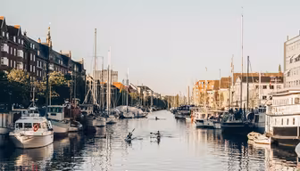 En fridfull kanalscen med flera små båtar som ligger till kaj längs kanterna och människor som paddlar kajak i vattnet. Kanalen kantas av tegelbyggnader och träd under en klarblå himmel.
