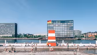 En solig dag vid en hamnpool med folk som badar och solar. En stor röd- och vitrandig ramp är framträdande, med moderna byggnader och en klarblå himmel i bakgrunden. Flaggor syns ovanpå rampen.