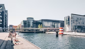 Människor solar på trädäck vid en kanal i ett tätortsområde. Moderna byggnader, inklusive en märkt "SØTORVET", ramar in scenen, med en liten bro som korsar vattnet. En randig boj syns i kanalen.