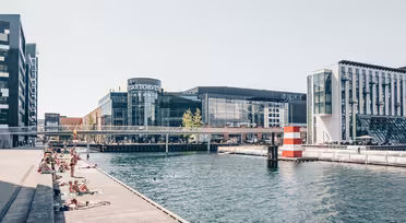 Människor solar på trädäck vid en kanal i ett tätortsområde. Moderna byggnader, inklusive en märkt "SØTORVET", ramar in scenen, med en liten bro som korsar vattnet. En randig boj syns i kanalen.
