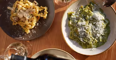 En bordsskiva med två tallrikar pasta: en med lockiga nudlar och den andra med grön ravioli, båda toppade med riven ost. Det finns vinglas fyllda med vitt vin och en hand som hälls upp från en mörk flaska.