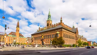 En livlig scen i Köpenhamns rådhus, med dess röda tegelarkitektur och gröna spiror under en delvis molnig himmel. Människor går i förgrunden nära en fontän, och den livliga gatan ger liv till den historiska miljön.