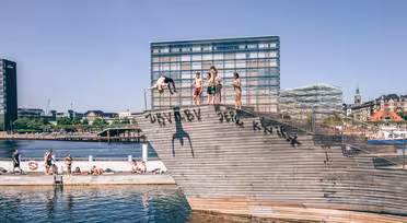 Människor är samlade på en träkonstruktion vid en strand, och några förbereder sig för att dyka i vattnet. Moderna byggnader och klar himmel finns i bakgrunden. Graffiti är synligt på strukturen.