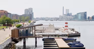 En scen vid vattnet med en modern träbyggnad, en dockad rad av små båtar och en industriell skyline med skorstenar och moderna byggnader i bakgrunden. Människor går längs en strandpromenad kantad av grönska.