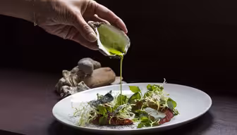 En hand häller grön vätska från ett ostronskal på en vit tallrik med diverse grönsaker, ätbara blommor och tunt skivat kött. Rätten är konstnärligt arrangerad, och ostronskal sitter i bakgrunden.