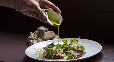 En hand häller grön vätska från ett ostronskal på en vit tallrik med diverse grönsaker, ätbara blommor och tunt skivat kött. Rätten är konstnärligt arrangerad, och ostronskal sitter i bakgrunden.