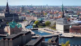 Flygfoto över Köpenhamn, som visar upp historisk och modern arkitektur med kanaler som skär genom staden. Framstående byggnader och kyrktorn reser sig ovanför, omgivna av träd och urban infrastruktur under en klarblå himmel.