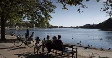 Två personer sitter på en bänk vid en sjö, omgivna av fåglar och cyklar. Sjön reflekterar den klarblå himlen, medan träd ger skugga. En person står nära en cykel och njuter av det fridfulla landskapet.