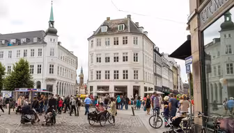 En livlig europeisk gatubild med människor som går, chattar och cyklar. Området har historiska flervåningsbyggnader med stora fönster. Ett café och butiker syns med ett torn i bakgrunden under en molnig himmel.