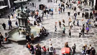 Ett livligt europeiskt torg med människor som går och samlas. Det finns en fontän till vänster och historiska byggnader med olika fasader kantar gatorna. Kullerstensmönster täcker marken och atmosfären är livlig och levande.