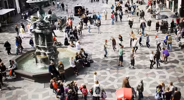 Ett livligt europeiskt torg med människor som går och samlas. Det finns en fontän till vänster och historiska byggnader med olika fasader kantar gatorna. Kullerstensmönster täcker marken och atmosfären är livlig och levande.