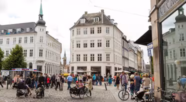 En livlig europeisk gatubild med människor som går och cyklar på kullerstenar. Omgivande byggnader är klassiska och utsmyckade, med en framstående byggnad med en spira. Kaféer och butiker kantar gatan, och flera träd ger grönska.