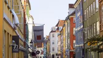 En levande gatubild i Köpenhamn med färgglada byggnader på båda sidor. En dansk flagga hänger bland byggnaderna. Fotgängare går längs kullerstensgatan medan bilar parkeras i närheten under en klarblå himmel.