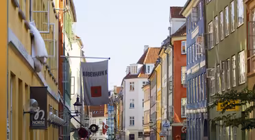 En levande gatubild i Köpenhamn med färgglada byggnader på båda sidor. En dansk flagga hänger bland byggnaderna. Fotgängare går längs kullerstensgatan medan bilar parkeras i närheten under en klarblå himmel.