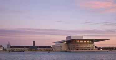 Bilden visar Köpenhamns operahus i solnedgången, reflekterat i den lugna hamnen. Byggnaden har en modern design med vidsträckta glasfönster och ett brett platt tak. Himlen är målad med nyanser av lila och rosa.