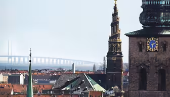 Stadsbild med ett kyrktorn med invecklade mönster och en klocka, byggnader med röda tak och en avlägsen bro som sträcker sig över vatten under en klar himmel.
