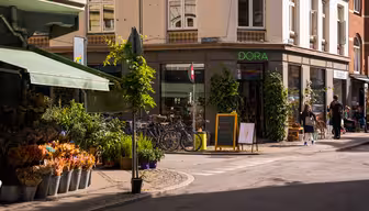Ett soligt gathörn med en blomsteraffär som visar färgglada buketter i hinkar. I närheten står cyklar parkerade utanför en butik som heter "DORA". En svart tavla med en glassteckning står vid entrén. Folk går längs trottoaren.