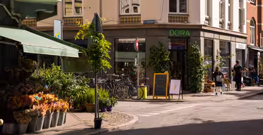 Ett soligt gathörn med en blomsteraffär som visar färgglada buketter i hinkar. I närheten står cyklar parkerade utanför en butik som heter "DORA". En svart tavla med en glassteckning står vid entrén. Folk går längs trottoaren.
