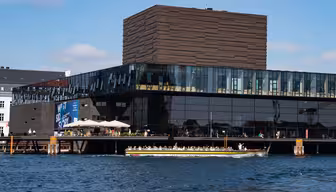 En modern byggnad med stora glasfönster sträcker sig över en strand, med en unik strukturerad brun toppsektion. En turbåt fylld med människor passerar framför på det blå vattnet under en klar himmel.