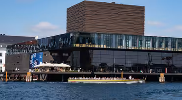 En modern byggnad med stora glasfönster sträcker sig över en strand, med en unik strukturerad brun toppsektion. En turbåt fylld med människor passerar framför på det blå vattnet under en klar himmel.