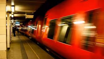 Ett rött tåg rusar genom en tunnelbanestation och skapar en rörelseoskärpa. Stationen har klinkergolv med gul taktil stenläggning. Taket är kantat med lysrör, och en person är synlig på avstånd.