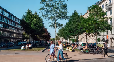 På ett livligt torg en solig dag kan folk koppla av och umgås. Två kvinnor går med en cykel, omgivna av träd och byggnader. Bilar står parkerade längs gatan och grupper av människor sitter tillsammans i bakgrunden.