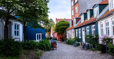 En pittoresk kullerstensgata kantad av färgglada hus med röda tak och gröna detaljer. Frodiga träd och buskar omger området, och cyklar står parkerade längs kanterna. Himlen är mulen, vilket ger en lugn atmosfär.