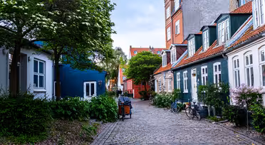 En pittoresk kullerstensgata kantad av färgglada hus med röda tak och gröna detaljer. Frodiga träd och buskar omger området, och cyklar står parkerade längs kanterna. Himlen är mulen, vilket ger en lugn atmosfär.