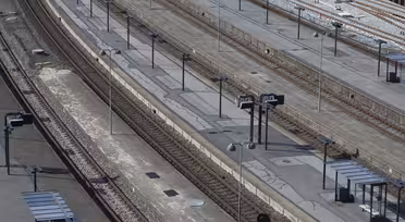 En flygbild över en tom tågstation med flera parallella spår och plattformar. Det finns små skyddsrum och skyltar på plattformarna, utan folk närvarande. Inställningen ser ljus och tydlig ut.