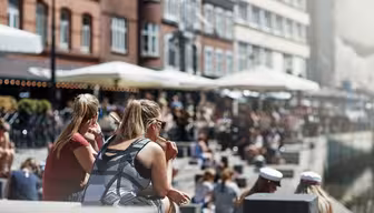 Människor sitter längs en kanal en solig dag, med tegelbyggnader och uteserveringar längs vattnet. Scenen är livlig, med individer som njuter av vädret, några bär solglasögon och andra i vita kepsar.