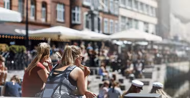 Människor sitter längs en kanal en solig dag, med tegelbyggnader och uteserveringar längs vattnet. Scenen är livlig, med individer som njuter av vädret, några bär solglasögon och andra i vita kepsar.