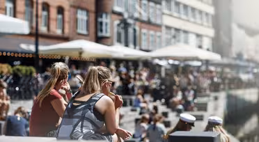 Människor sitter längs en kanal en solig dag, med tegelbyggnader och uteserveringar längs vattnet. Scenen är livlig, med individer som njuter av vädret, några bär solglasögon och andra i vita kepsar.