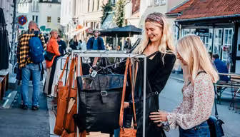 Två kvinnor ler medan de tittar på läderväskor på ett displayställ utomhus. En gata med andra människor och butiker ligger i bakgrunden. De verkar njuta av en solig dag med shopping i en charmig stadsdel.