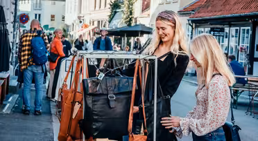 Två kvinnor ler medan de tittar på läderväskor på ett displayställ utomhus. En gata med andra människor och butiker ligger i bakgrunden. De verkar njuta av en solig dag med shopping i en charmig stadsdel.