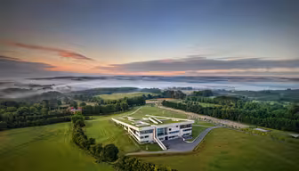 Flygfoto över en modern byggnad omgiven av vidsträckta gröna fält och träd vid soluppgången. Dimma rullar över avlägsna kullar under en delvis molnig himmel med varma nyanser. Byggnaden har en unik kantig design och kantas av en slingrande uppfart.