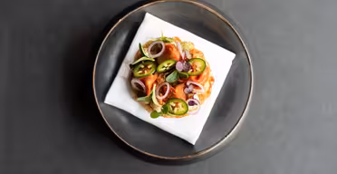 En pläterad maträtt med en välsmakande bakelse toppad med skivade jalapeños, lökringar och färska örter, serverad på en vit servett i ett runt, mörkt fat mot en mörk bakgrund.