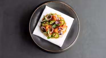 En pläterad maträtt med en välsmakande bakelse toppad med skivade jalapeños, lökringar och färska örter, serverad på en vit servett i ett runt, mörkt fat mot en mörk bakgrund.