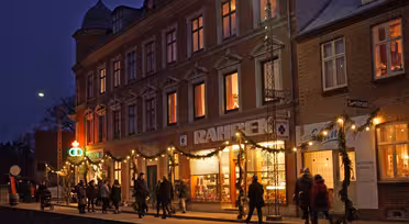 En grupp människor går längs en festlig gata på kvällen. Den stora byggnaden, prydd med semesterbelysning och girlanger, har butiker med upplysta fönster, inklusive ett bryggeri och snackbar. Himlen är mörk och scenen är varmt upplyst.