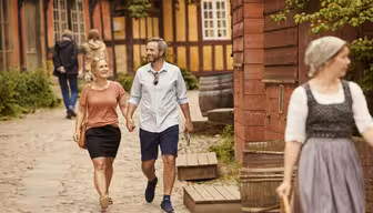 Ett leende par går hand i hand på en kullerstensgata med traditionella byggnader i bakgrunden. En person i traditionella kläder går i förgrunden. Andra människor syns längre fram på stigen.