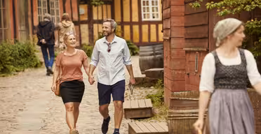 Ett leende par går hand i hand på en kullerstensgata med traditionella byggnader i bakgrunden. En person i traditionella kläder går i förgrunden. Andra människor syns längre fram på stigen.