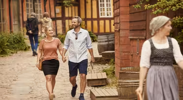 Ett leende par går hand i hand på en kullerstensgata med traditionella byggnader i bakgrunden. En person i traditionella kläder går i förgrunden. Andra människor syns längre fram på stigen.