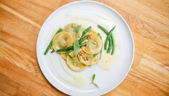 En vit tallrik med ravioli toppad med pinjenötter och garnerad med färsk basilika. Gröna bönor är ordnade bredvid, med en blek sås som droppas konstnärligt. Plattan ställs på en träyta.