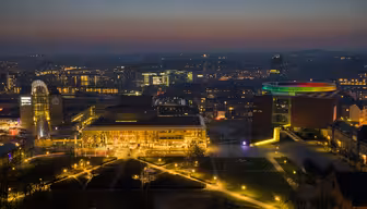 Flygfoto nattvy av en stadsbild med upplysta byggnader och gator. Himlen är en gradient av blått och orange. En framstående byggnad har en livfull, mångfärgad cirkulär struktur på taket, vilket lägger till en färgklick till scenen.