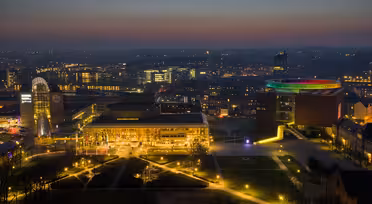 Flygfoto nattvy av en stadsbild med upplysta byggnader och gator. Himlen är en gradient av blått och orange. En framstående byggnad har en livfull, mångfärgad cirkulär struktur på taket, vilket lägger till en färgklick till scenen.