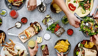 Flygfoto över ett bord med händer som sträcker sig efter olika rätter, inklusive tacos, sallad, pommes frites, chips och dipp. Drycker och garnityr som limeklyftor finns också, vilket skapar en livlig och hektisk matscen.