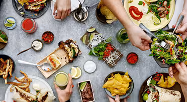 Flygfoto över ett bord med händer som sträcker sig efter olika rätter, inklusive tacos, sallad, pommes frites, chips och dipp. Drycker och garnityr som limeklyftor finns också, vilket skapar en livlig och hektisk matscen.